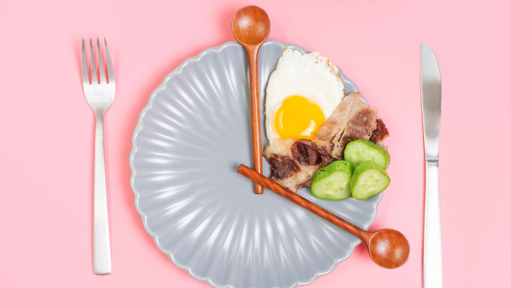 Clock shaped wooden spoons on plate with food on pink background top view, intermittent fasting concept.