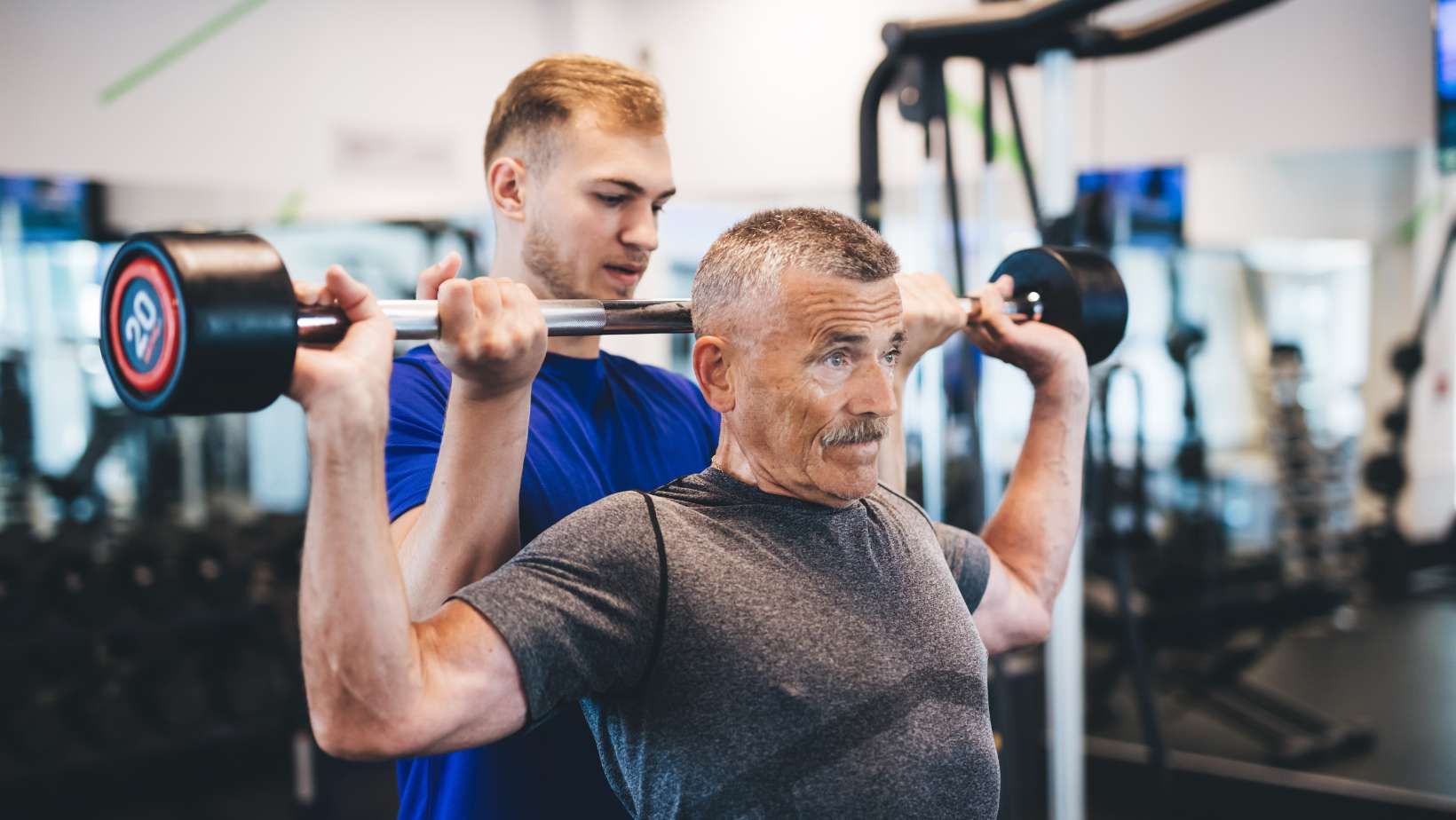 Senior Man Lifting Weights with Help of Gym Assistant